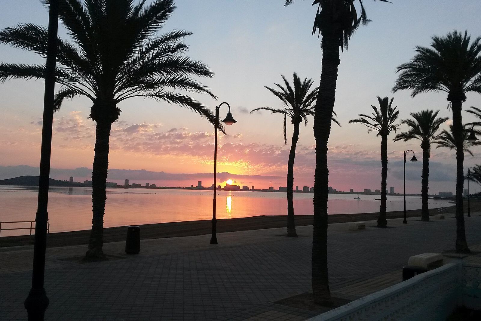 Päikesetõus Mar Menori kohal, vaadatuna laguuniäärselt kiviplaatidega promenaadilt: palmide ja laternapostide siluetid, roosa taevas ja peegelsile vesi.