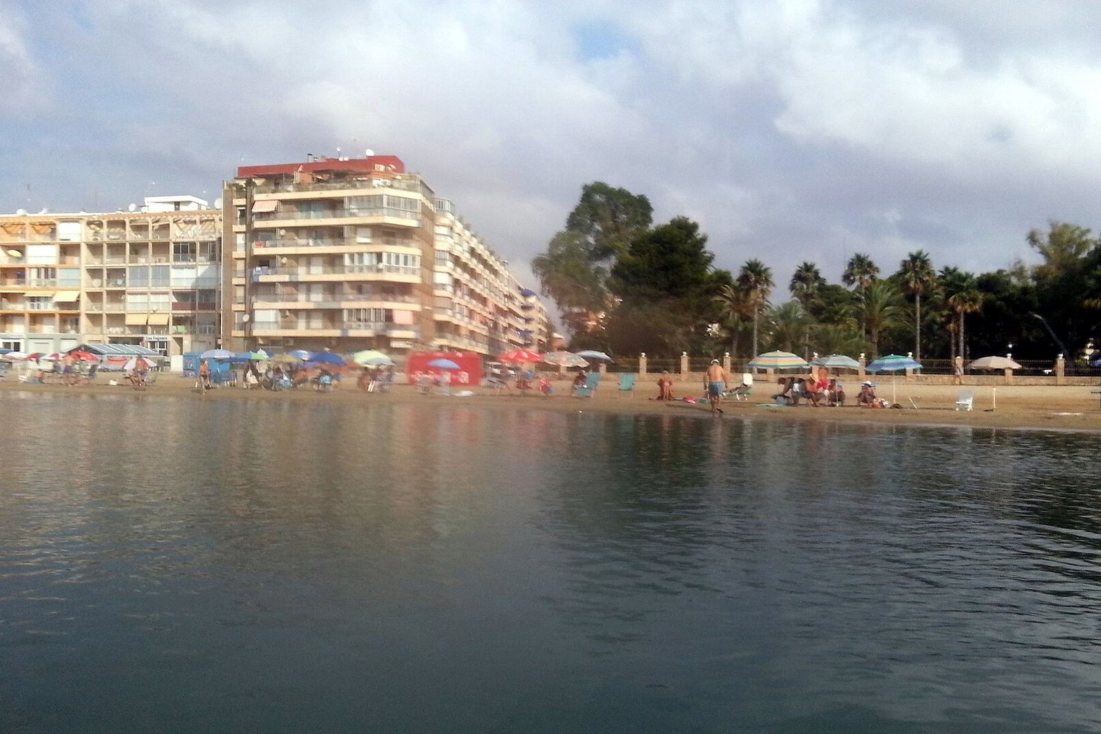 Torrevieja linnarand veelt vaadatuna, värvilised päikesevarjud ja päevitajad liival, taga kõrged kortermajad, palmirida ja pilvine taevas